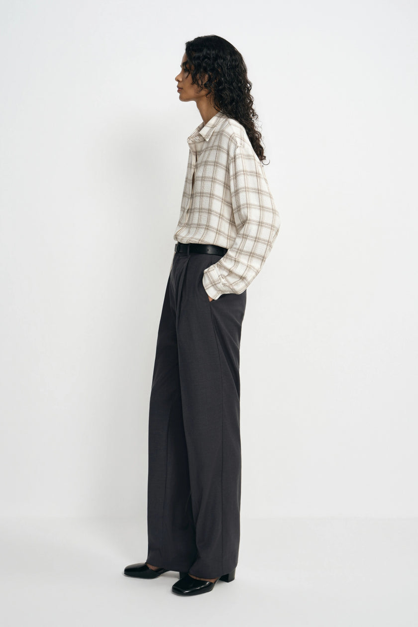 A model is shown from a side profile wearing a beige and white plaid button-down shirt tucked into dark charcoal gray wide-leg trousers, paired with black shoes, against a plain white studio background.