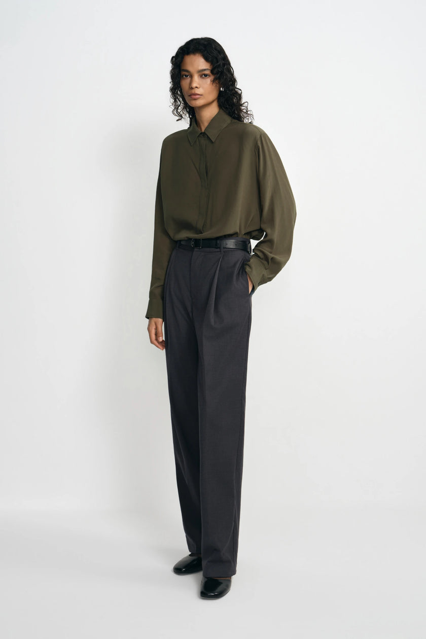 A model wearing dark grey pleat-front wide-leg trousers in a silky wool blend, pairing an olive shirt with a black belt and black loafers.