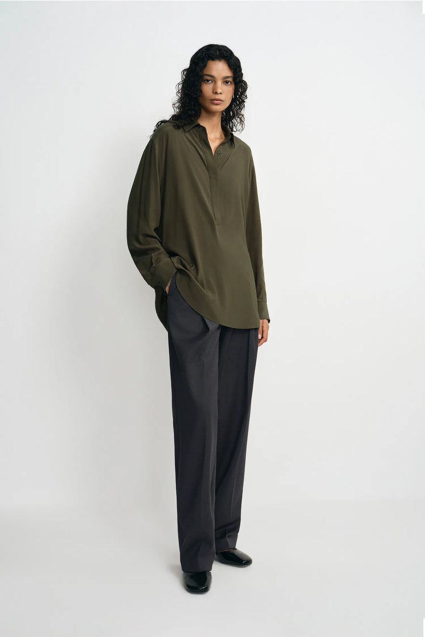 A model wearing dark grey pleat-front wide-leg trousers in a silky wool blend, pairing an oversized olive shirt and black loafers.