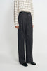 A model wearing dark grey pleat-front wide-leg trousers in a silky wool blend, close-up front view showing the pleat detail and belt loops, pairing a checked shirt and black belt.