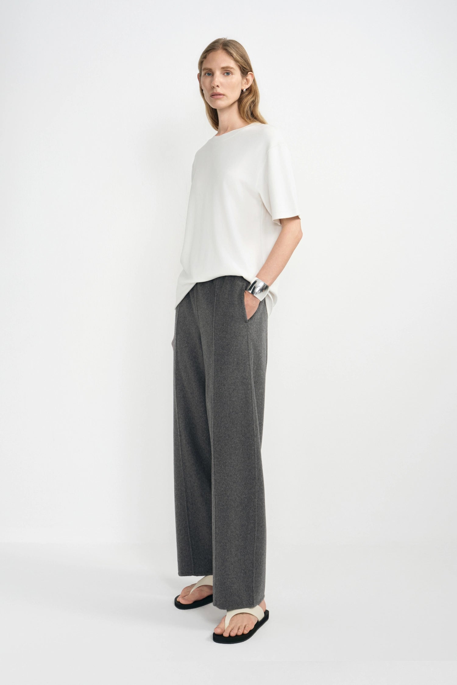A woman modeling a white short-sleeve top and dark gray wide-leg trousers, standing against a plain white studio background.