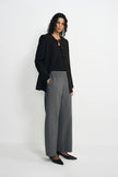 A model stands against a plain light gray studio background wearing a black cropped blazer over a black top, paired with high-waisted wide-leg gray trousers and black pointed flats.