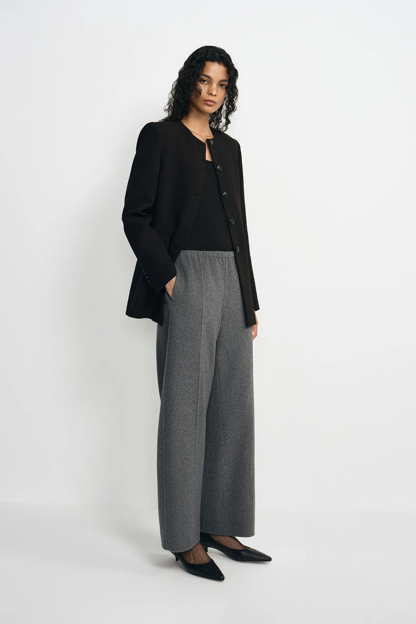 A model stands against a plain light gray studio background wearing a black cropped blazer over a black top, paired with high-waisted wide-leg gray trousers and black pointed flats.
