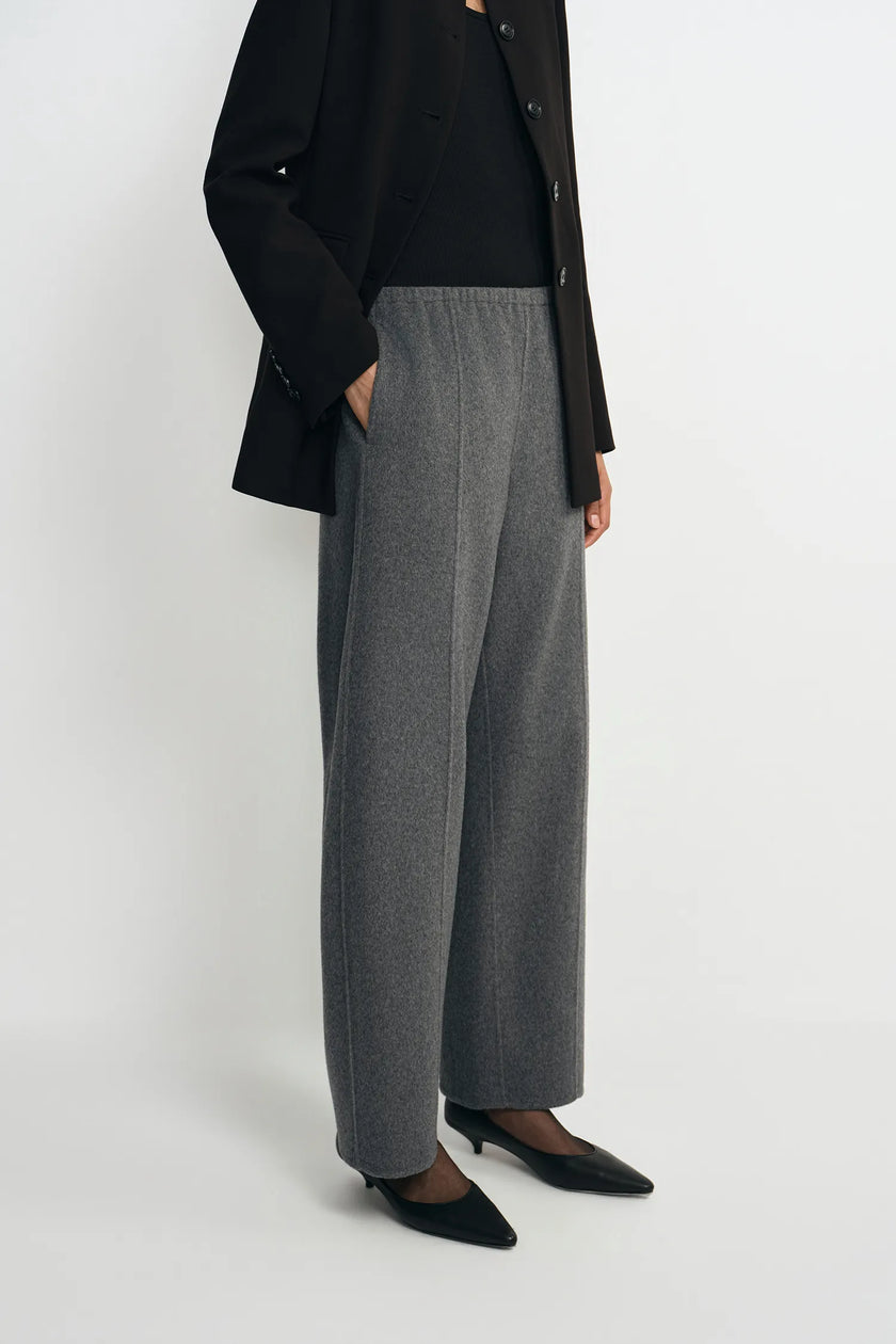 A model wearing a pair of wide-leg gray wool trousers, styled with a black blazer, black top, and black pointed-toe heels against a plain light gray studio background.