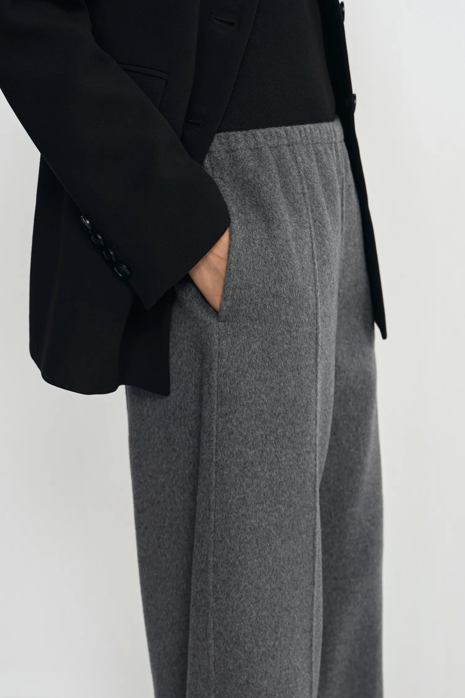 A close-up of a person wearing wide-leg gray trousers with a black blazer and black top, standing against a plain light gray background.