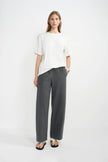 A woman stands against a plain light gray studio background wearing a loose white short-sleeve top, dark gray wide-leg trousers, and black sandals.