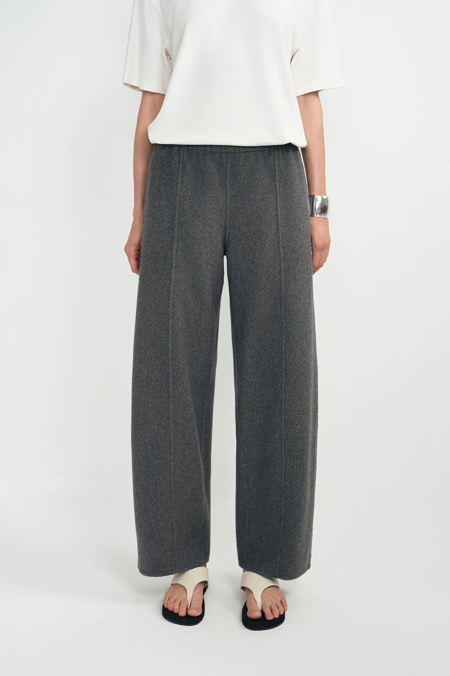 A model wearing dark gray wide-leg wool trousers, styled with a white short-sleeve top and black-and-white sandals against a plain white studio background.