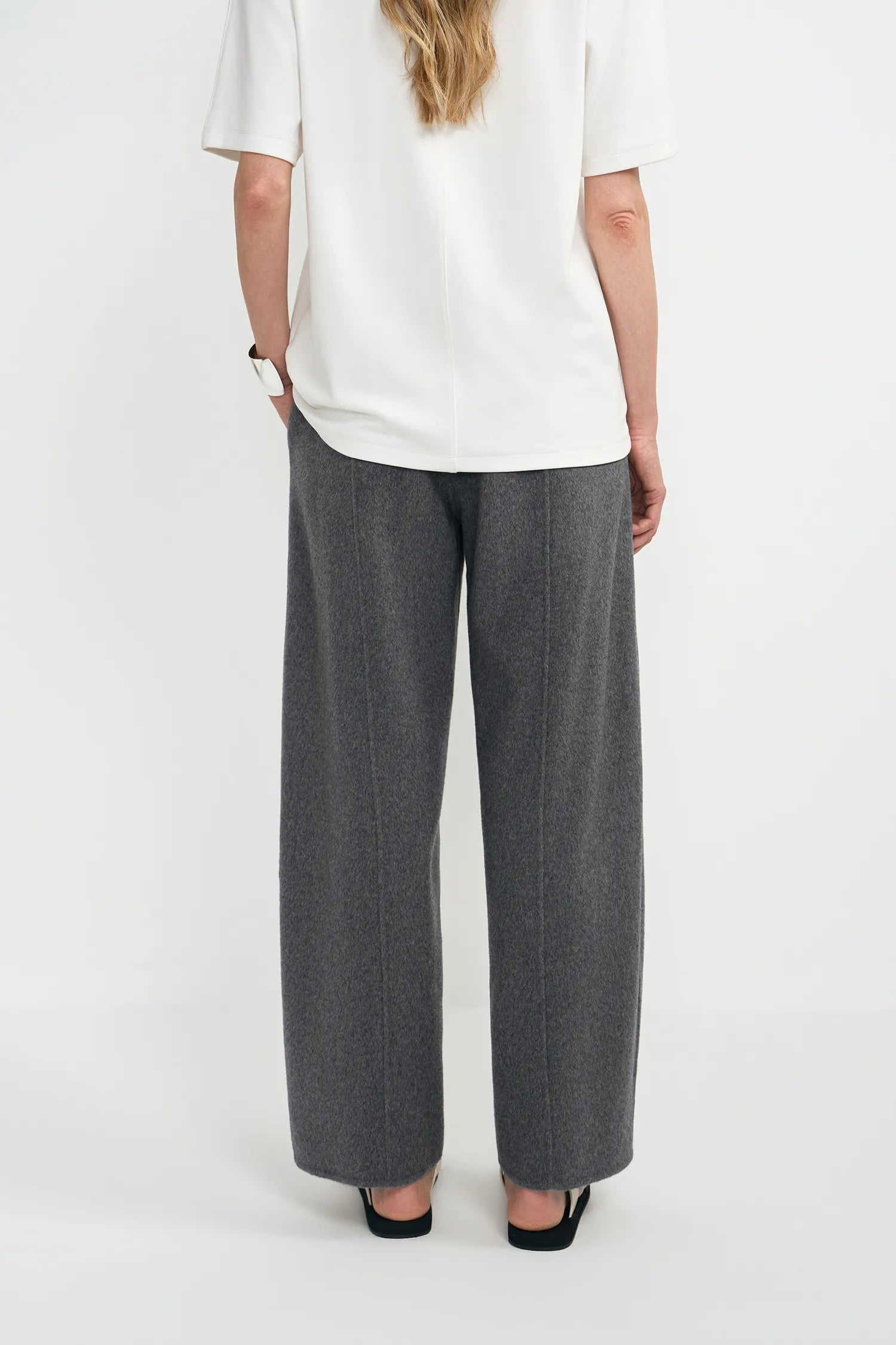 A model shown from the back wearing wide-leg gray trousers, a white short-sleeve top, and black flat shoes against a plain white studio background.