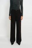 A model wearing dark brown wide-leg corduroy trousers, styled with a dark gray long-sleeve top and black pointed-toe heels, standing against a plain light gray studio background.
