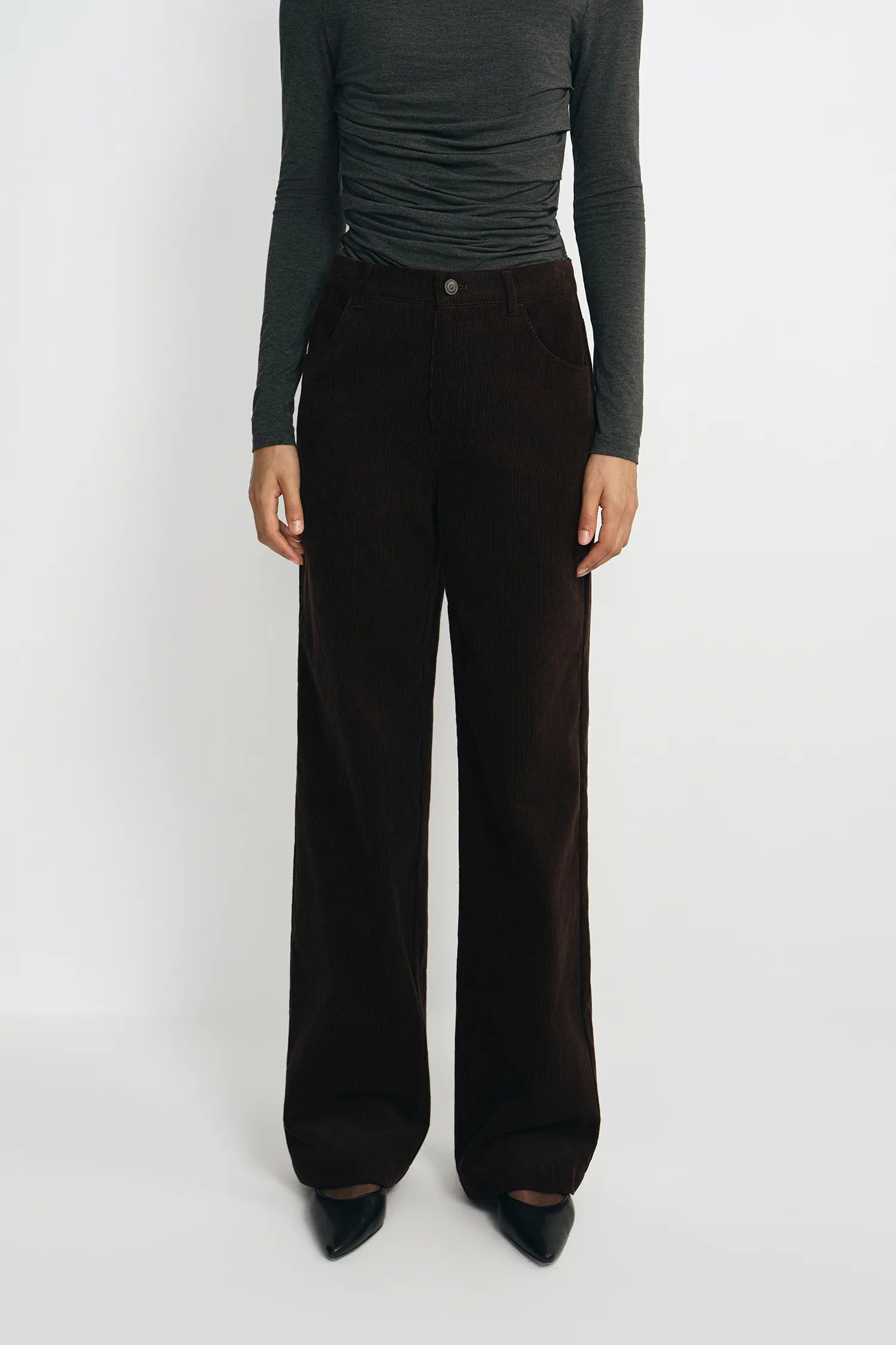 A model wearing dark brown wide-leg corduroy trousers, styled with a dark gray long-sleeve top and black pointed-toe heels, standing against a plain light gray studio background.