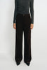 A model wearing dark brown wide-leg corduroy trousers, styled with a dark gray long-sleeve top and black pointed-toe heels, standing against a plain light gray studio background.