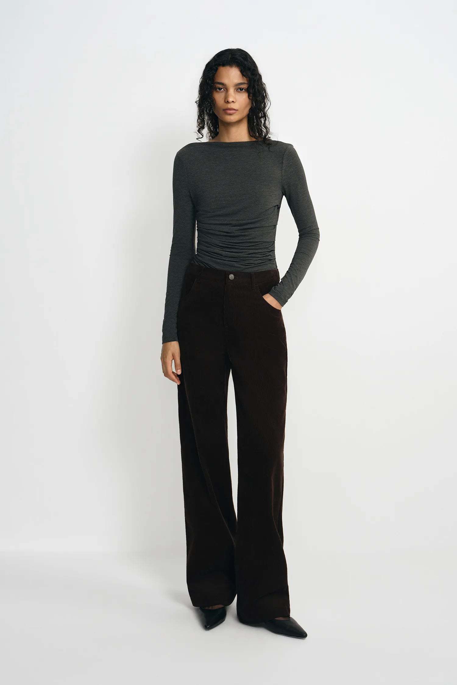 A model stands against a plain white studio background wearing a dark gray long-sleeve top and dark brown wide-leg trousers.