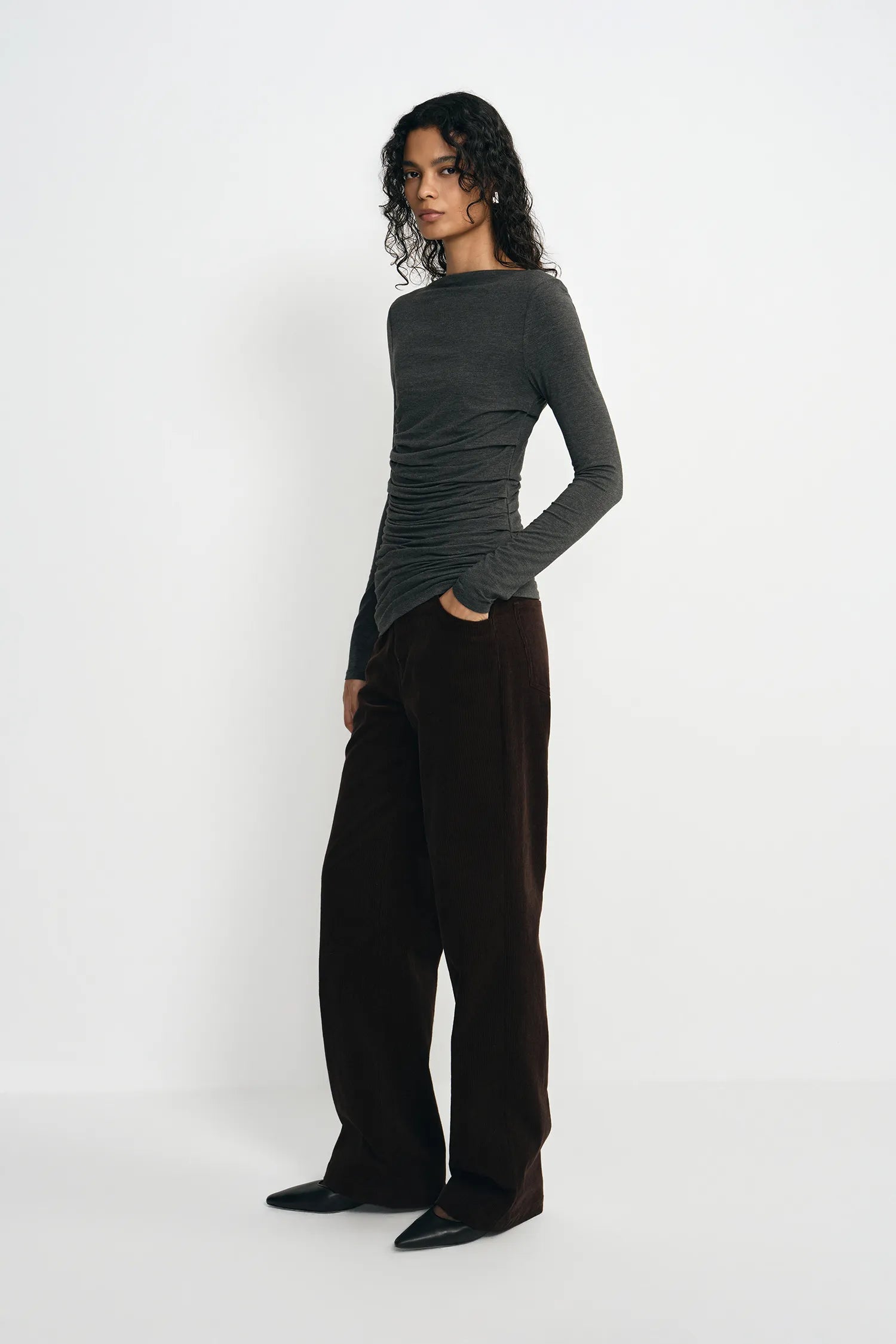 A model stands in profile wearing a dark charcoal gray long-sleeve ruched top and dark brown wide-leg trousers against a plain white studio background.