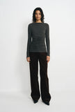 A model stands against a plain white studio background wearing a dark charcoal gray long-sleeve top with ruching and dark brown wide-leg trousers.