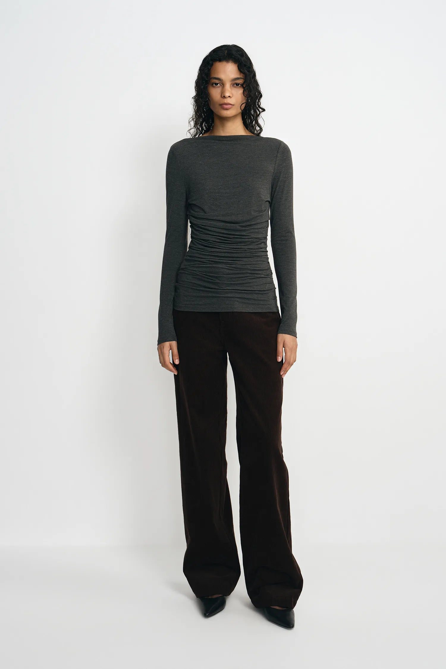 A model stands against a plain white studio background wearing a dark charcoal gray long-sleeve top with ruching and dark brown wide-leg trousers.