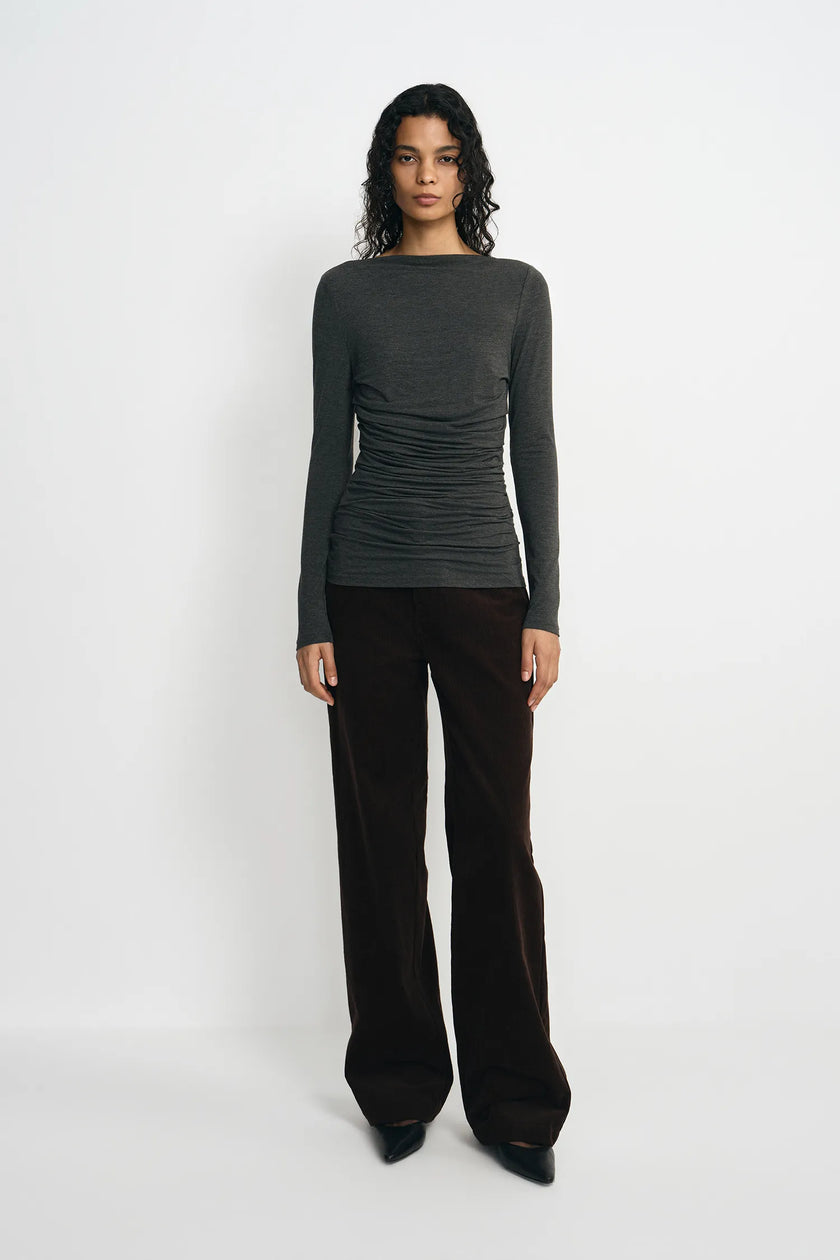 A model stands against a plain white studio background wearing a dark charcoal gray long-sleeve top with ruching and dark brown wide-leg trousers.