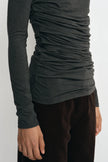 A close-up of a person wearing a dark charcoal gray long-sleeve top with ruched side detailing and dark brown pants, shown against a plain light background.