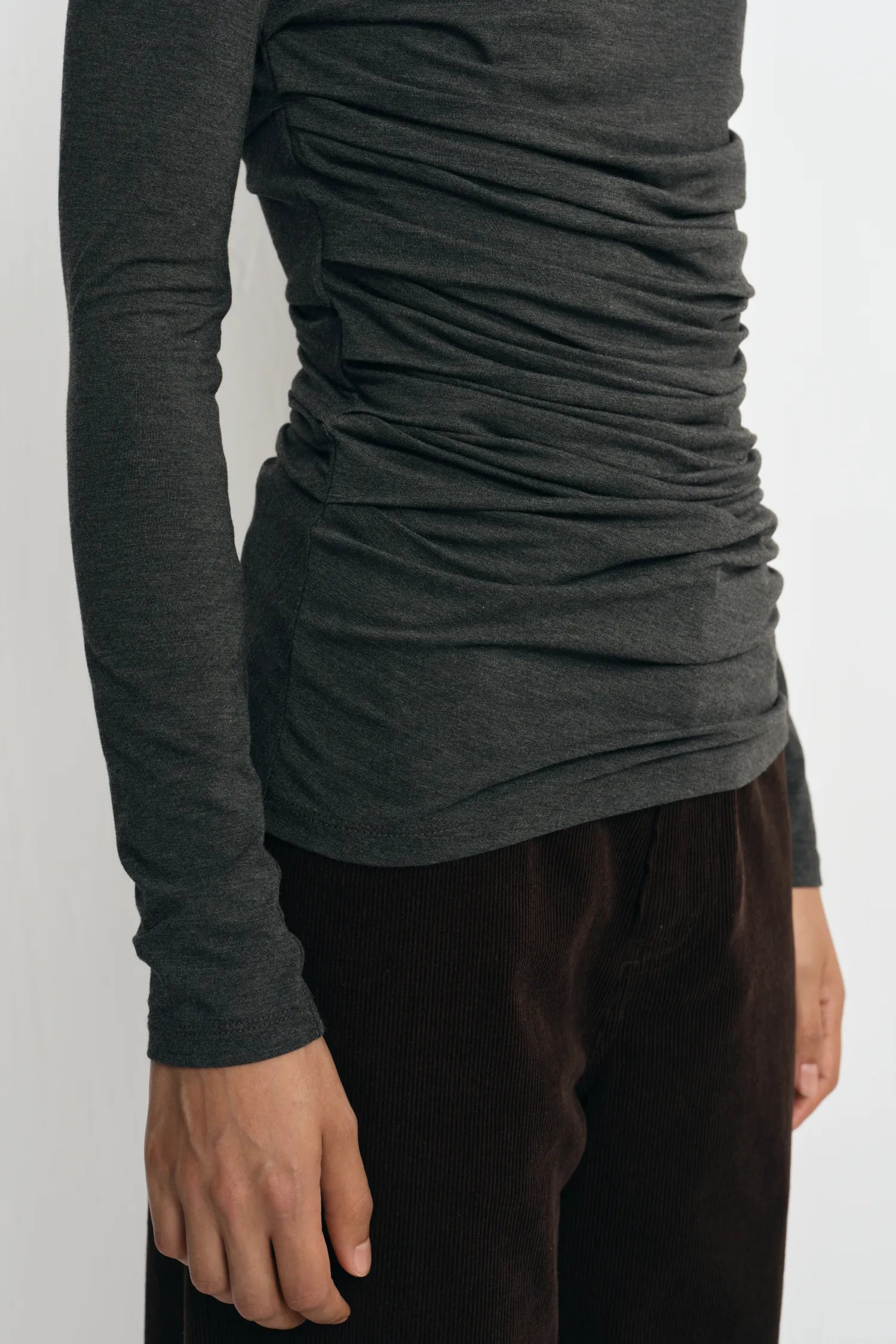 A close-up of a person wearing a dark charcoal gray long-sleeve top with ruched side detailing and dark brown pants, shown against a plain light background.