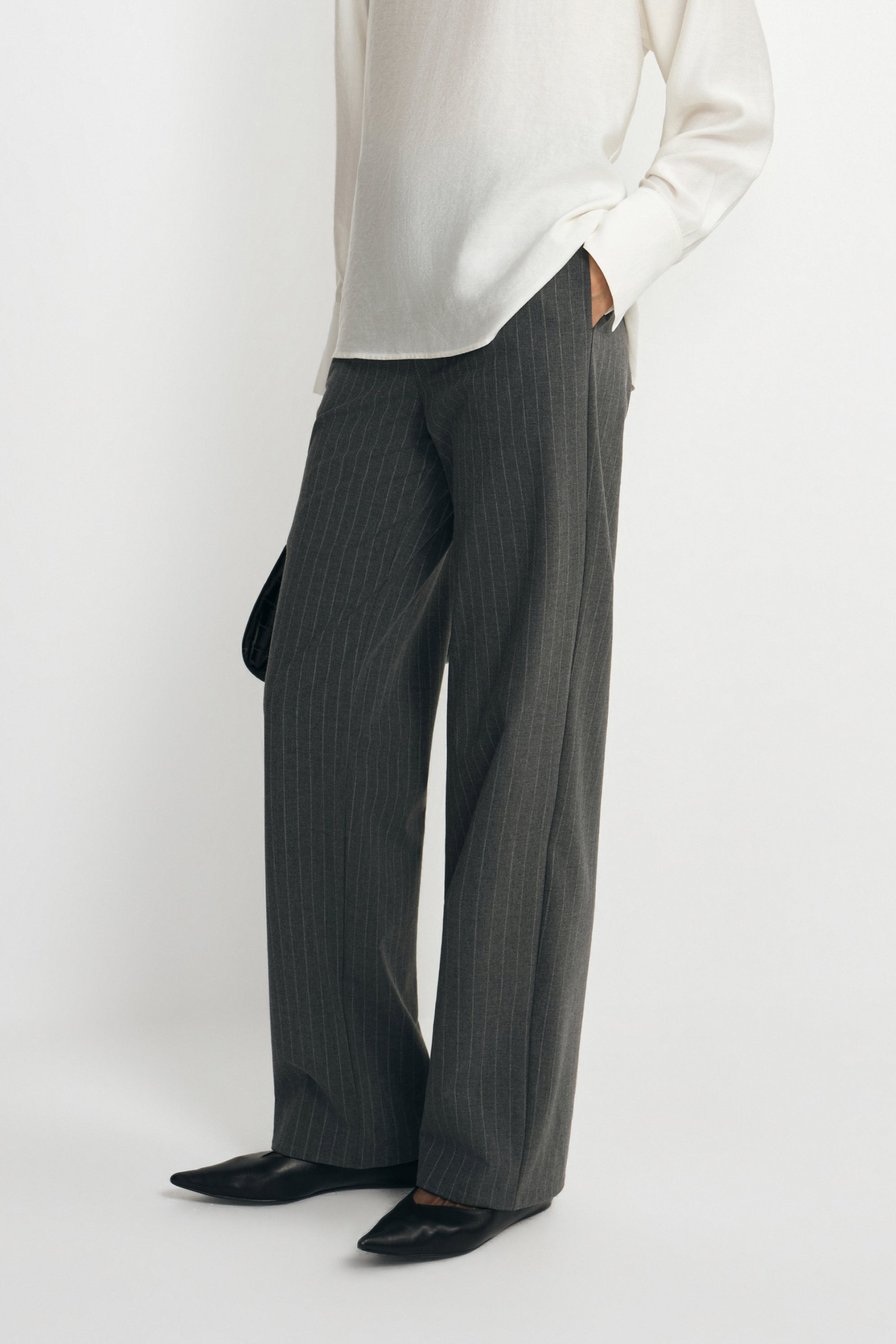 Close-up of charcoal trousers on a model, showing fine vertical stripes and straight legs extending over black pointed shoes beneath a white blouse.