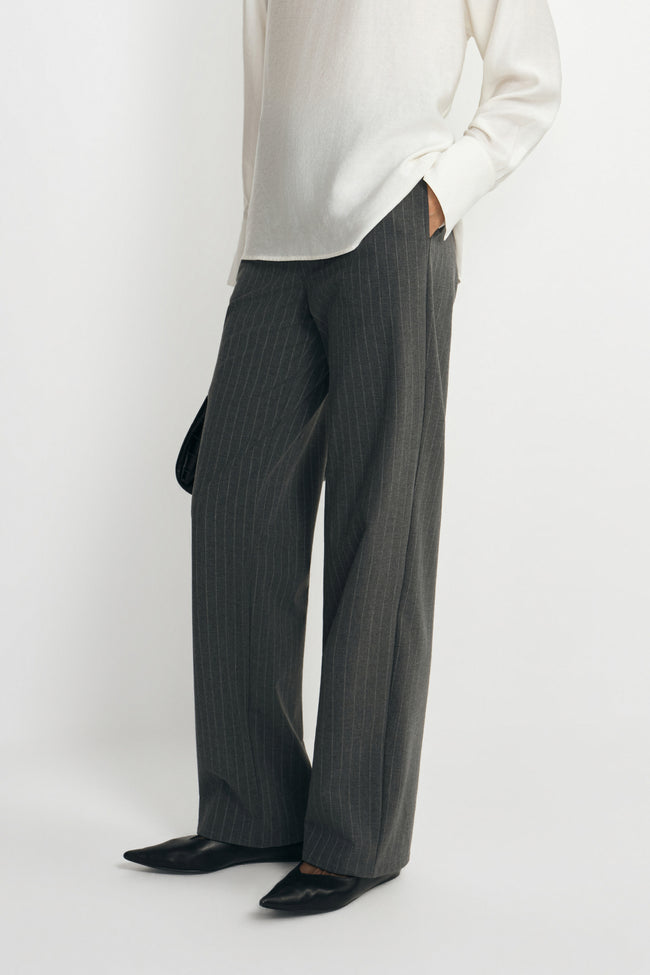Close-up of charcoal trousers on a model, showing fine vertical stripes and straight legs extending over black pointed shoes beneath a white blouse.