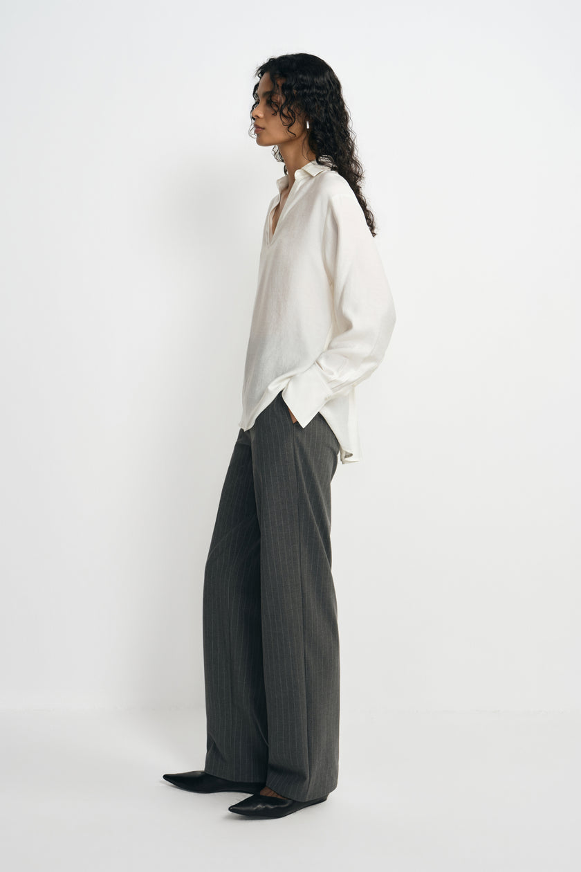 Side view of a model wearing charcoal pinstriped trousers with long straight legs, styled with an untucked white blouse and black pointed shoes.