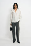 A model wearing charcoal pinstriped trousers with long straight legs, paired with an untucked white blouse, black pointed shoes and a black handbag.