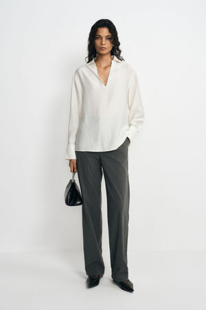 A model wearing charcoal pinstriped trousers with long straight legs, paired with an untucked white blouse, black pointed shoes and a black handbag.