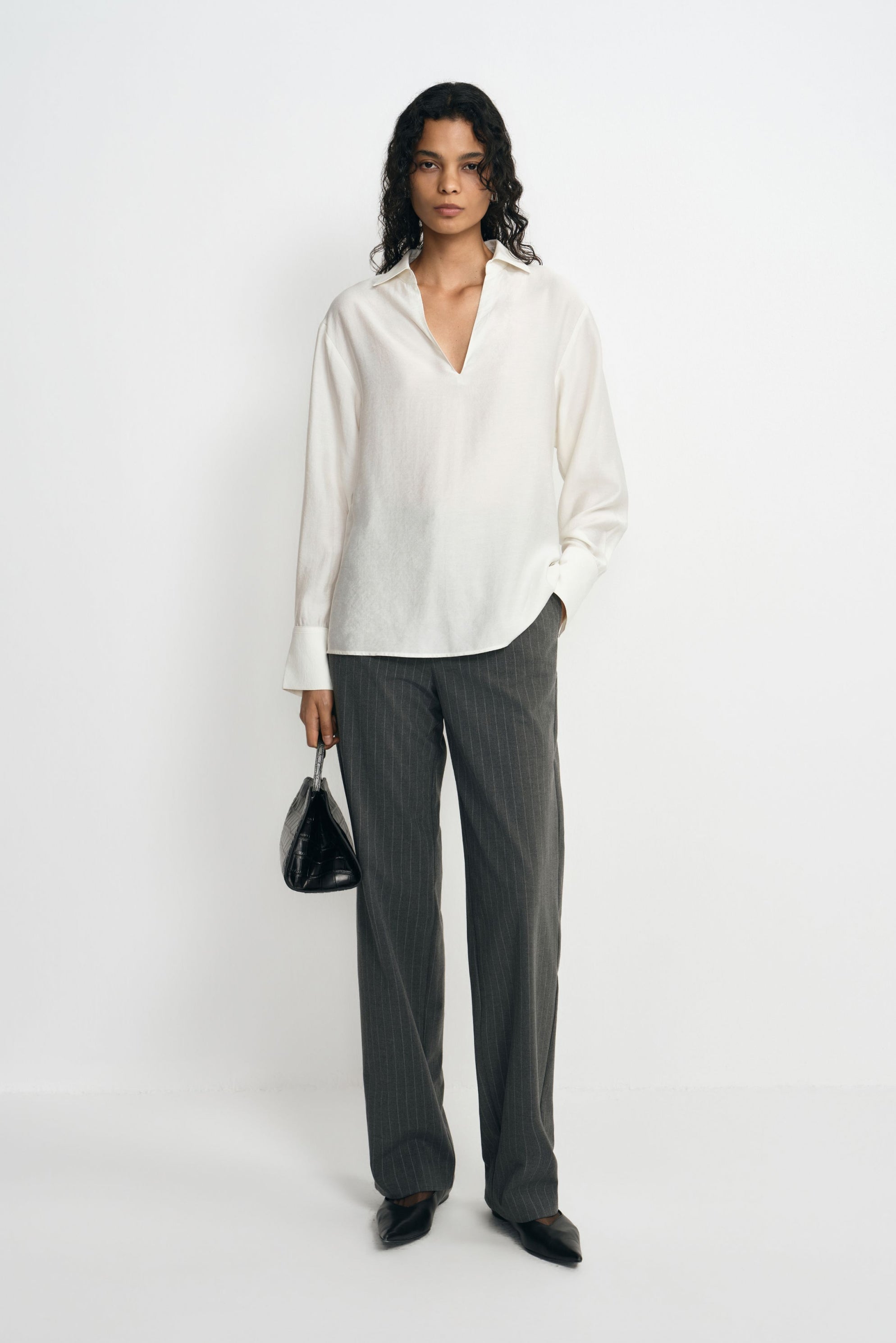 A model wearing charcoal pinstriped trousers with long straight legs, paired with an untucked white blouse, black pointed shoes and a black handbag.