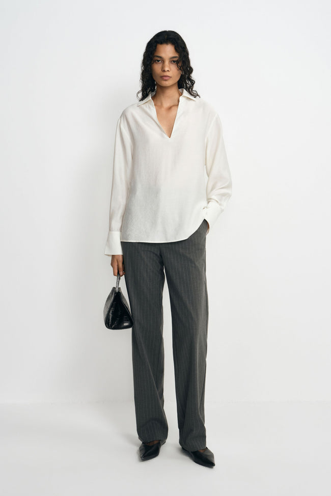 A model wearing charcoal pinstriped trousers with long straight legs, paired with an untucked white blouse, black pointed shoes and a black handbag.