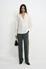 A model wearing charcoal pinstriped trousers with long straight legs, paired with an untucked white blouse, black pointed shoes and a black handbag.