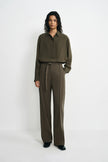 A model stands against a plain white studio background wearing an olive green long-sleeve button-up shirt and matching olive green wide-leg trousers with a tie belt.