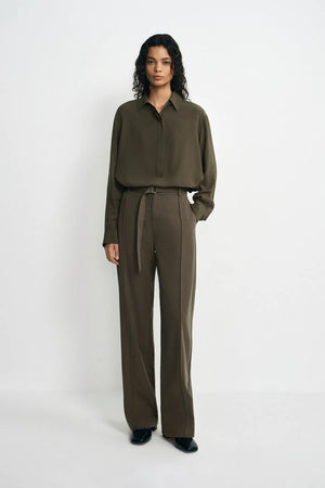 A model stands against a plain white studio background wearing an olive green long-sleeve button-up shirt and matching olive green wide-leg trousers with a tie belt.