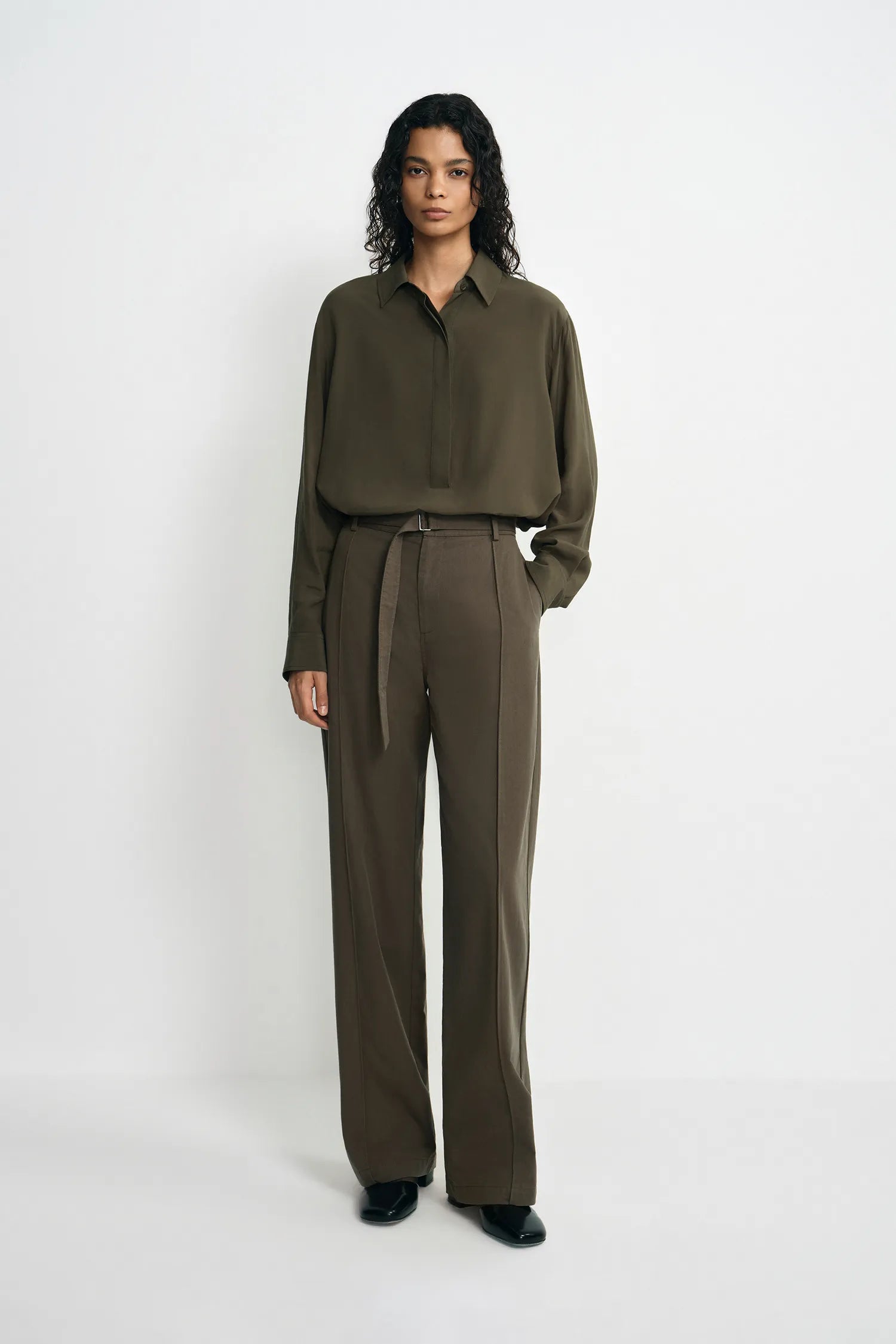 A model stands against a plain white studio background wearing an olive green long-sleeve button-up shirt and matching olive green wide-leg trousers with a tie belt.