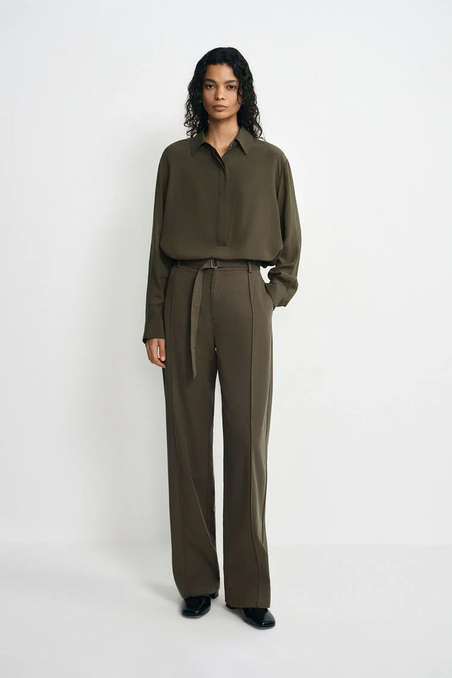 A model stands against a plain white studio background wearing an olive green long-sleeve button-up shirt and matching olive green wide-leg trousers with a tie belt.
