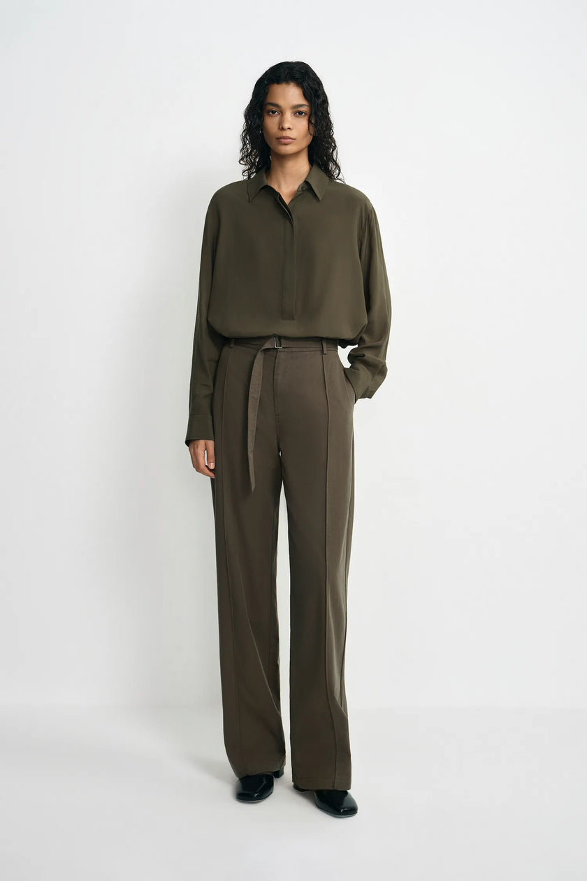 A model stands against a plain white studio background wearing an olive green long-sleeve button-up shirt and matching olive green wide-leg trousers with a tie belt.