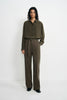 A model stands against a plain white studio background wearing an olive green long-sleeve button-up shirt and matching olive green wide-leg trousers with a tie belt.