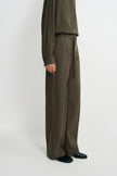 A model is shown from the waist down wearing high-waisted, wide-leg trousers in a dark olive brown color, paired with a matching long-sleeve top and black shoes, against a plain light gray studio background.