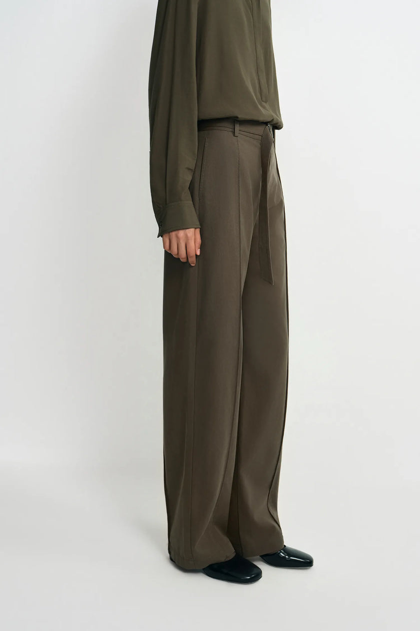 A model is shown from the waist down wearing high-waisted, wide-leg trousers in a dark olive brown color, paired with a matching long-sleeve top and black shoes, against a plain light gray studio background.