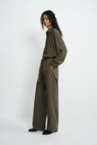 A model stands in profile wearing an olive green long-sleeve collared shirt and matching wide-leg trousers, paired with black shoes, against a plain light gray studio background.