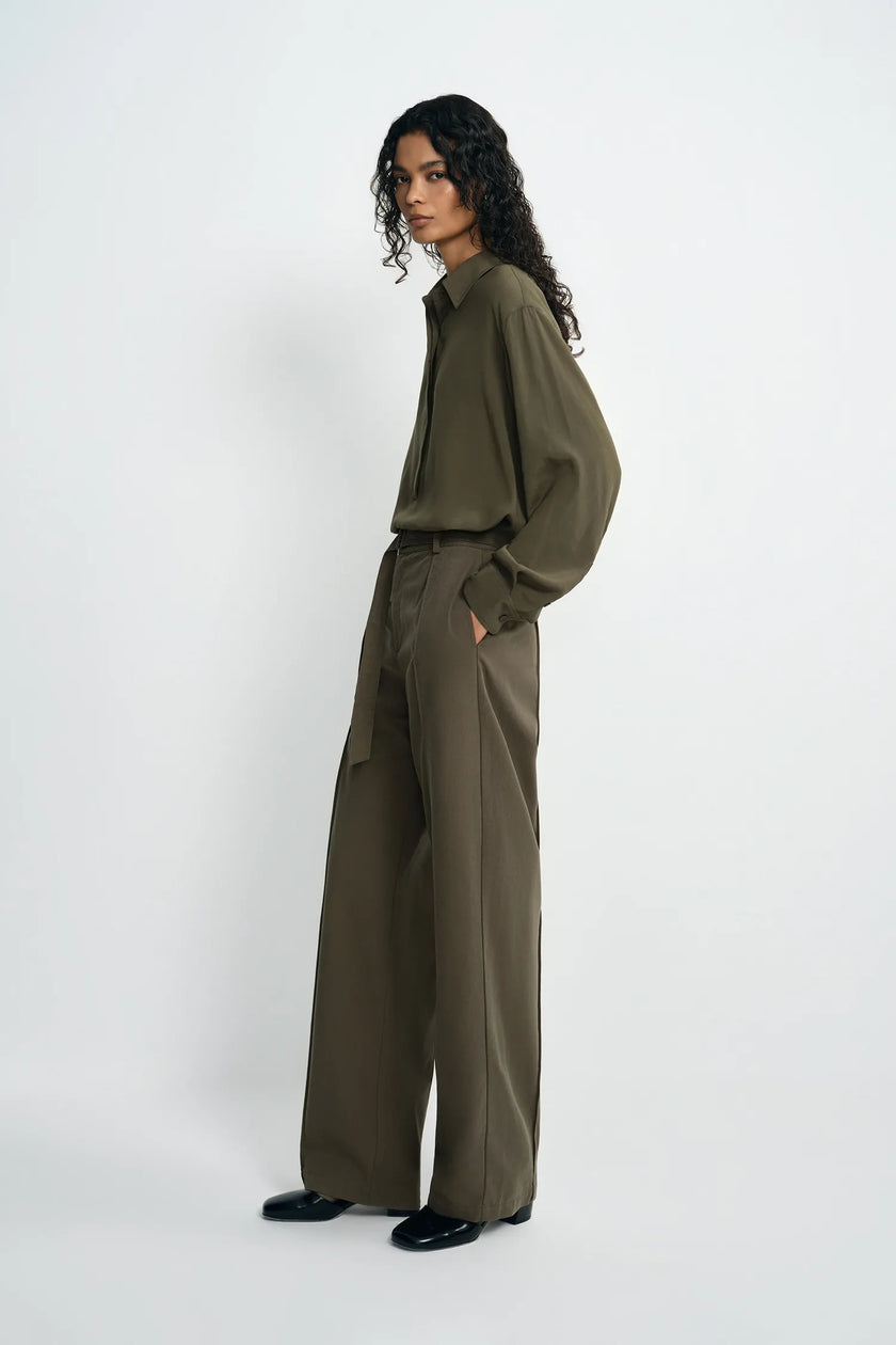 A model stands in profile wearing an olive green long-sleeve collared shirt and matching wide-leg trousers, paired with black shoes, against a plain light gray studio background.