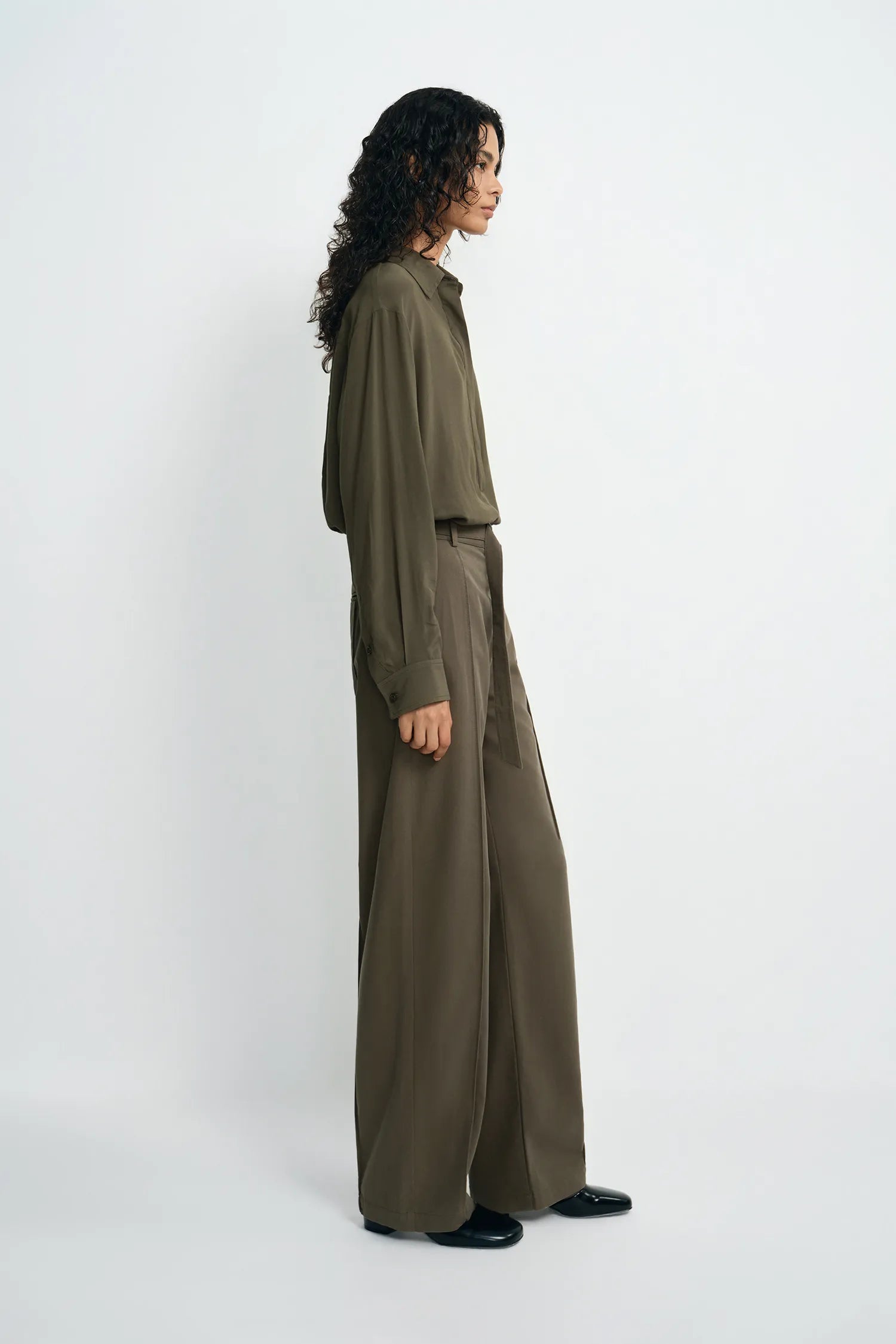 A model is shown from the side wearing a matching olive green long-sleeve button-up shirt and wide-leg trousers set, paired with black shoes, against a plain light gray studio background.