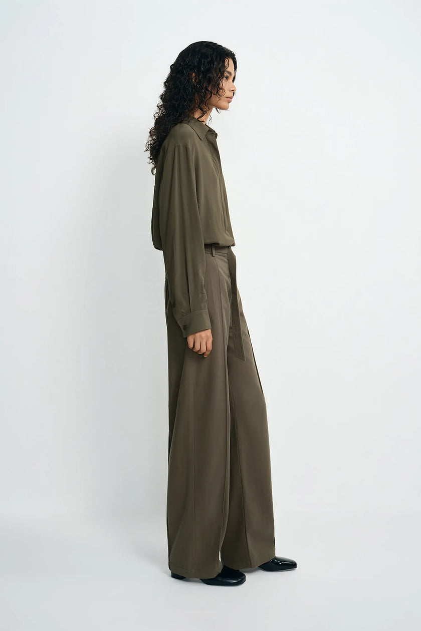 A model is shown from the side wearing a matching olive green long-sleeve button-up shirt and wide-leg trousers set, paired with black shoes, against a plain light gray studio background.