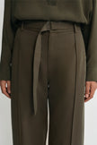 A close-up of a model wearing high-waisted olive green trousers with a matching fabric tie belt and a matching long-sleeve top, shown against a plain light gray background.