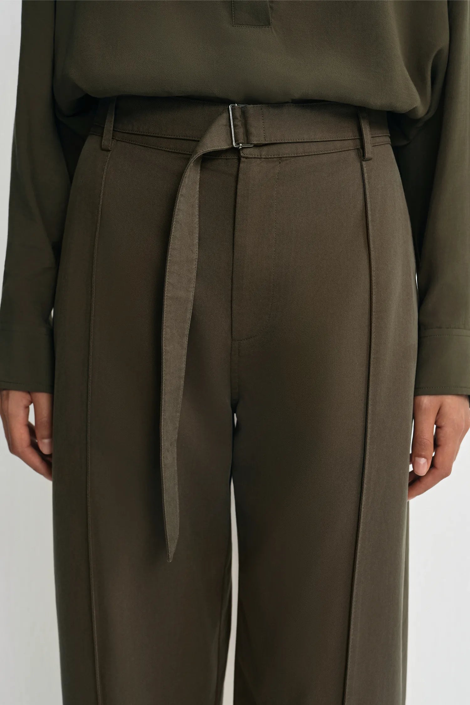 A close-up of a model wearing high-waisted olive green trousers with a matching fabric tie belt and a matching long-sleeve top, shown against a plain light gray background.