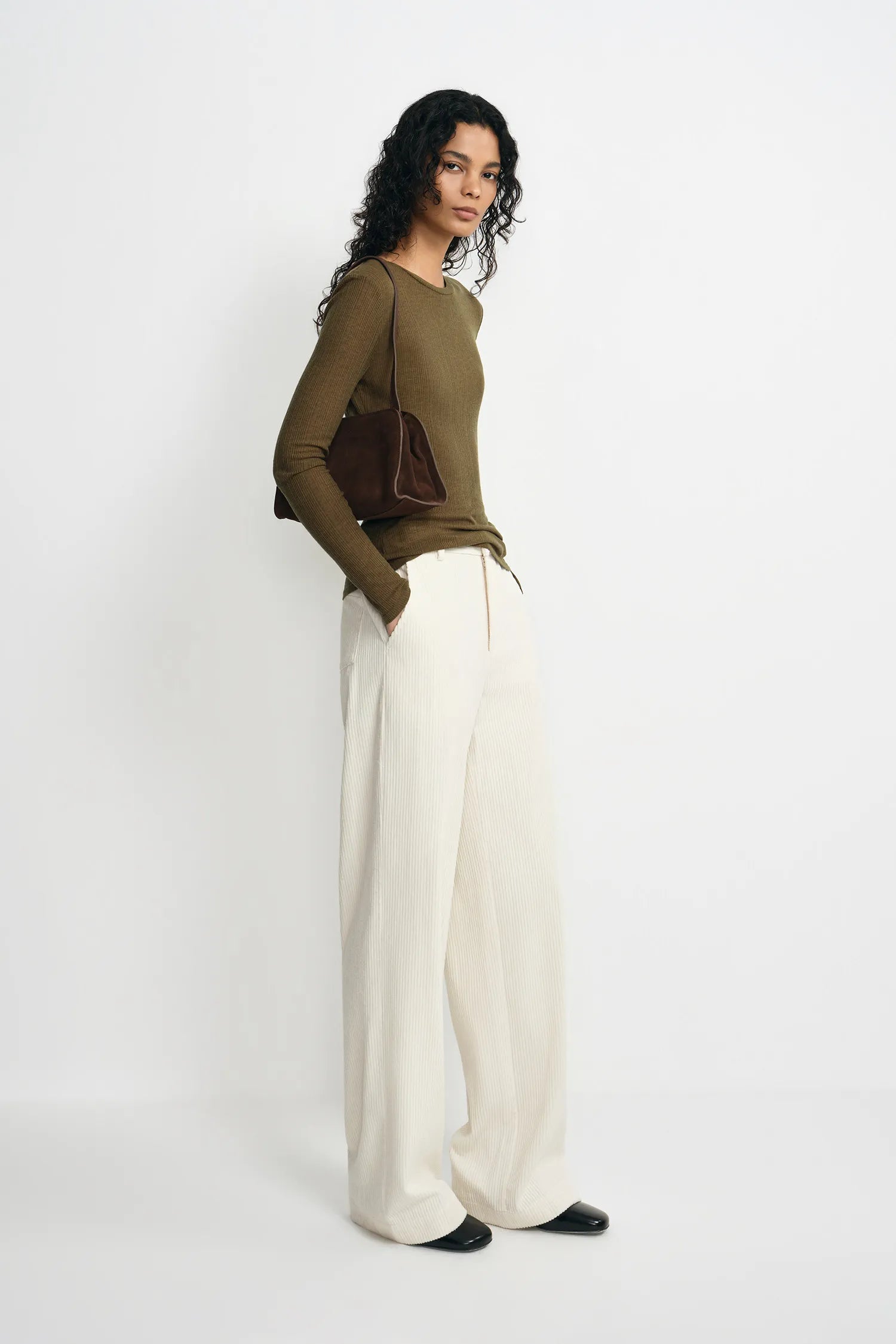 A woman stands in profile wearing a fitted olive green long-sleeve top, wide-leg cream trousers, and black shoes, with a dark brown shoulder bag against a plain light gray studio background.