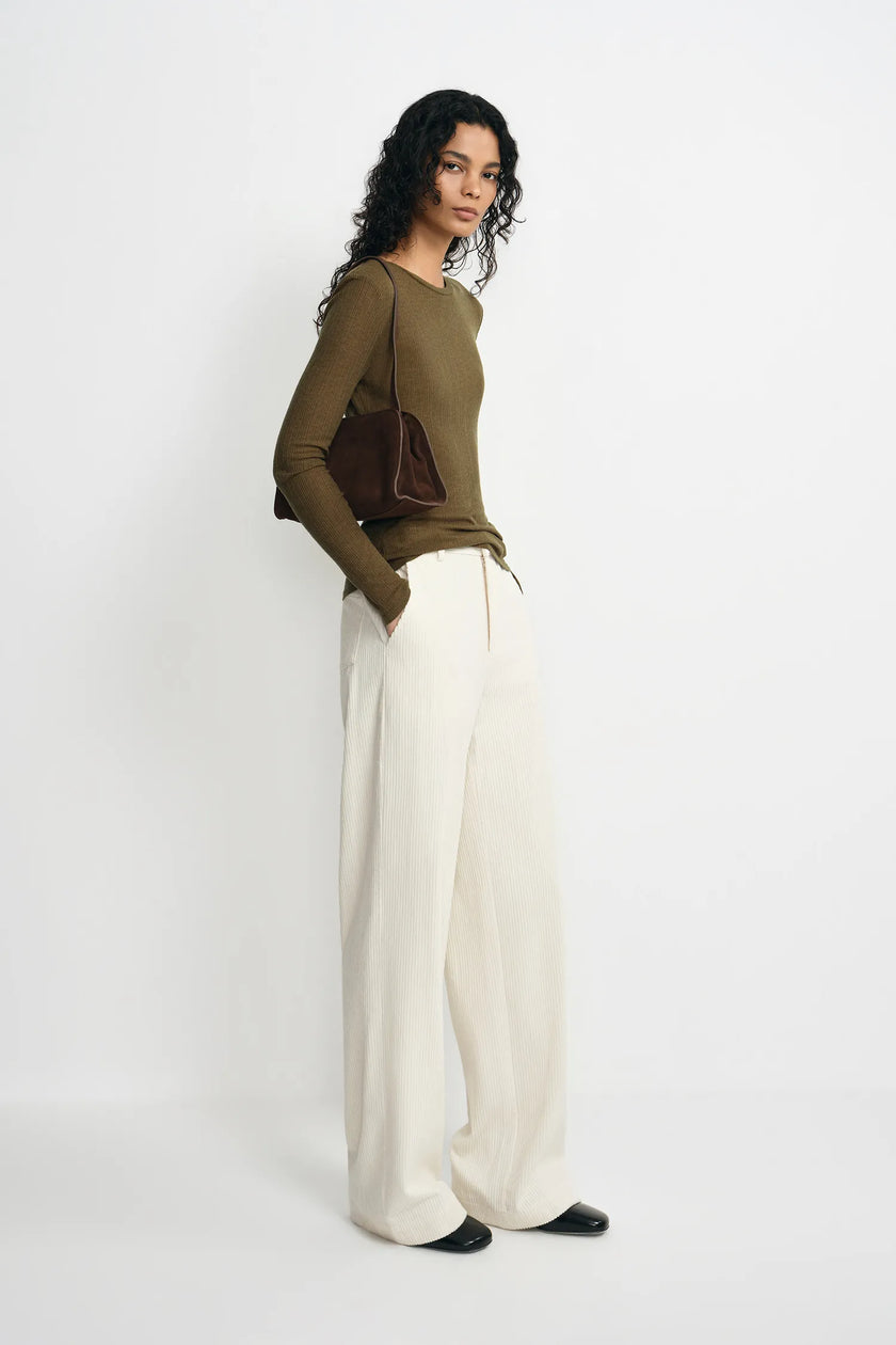 A woman stands in profile wearing a fitted olive green long-sleeve top, wide-leg cream trousers, and black shoes, with a dark brown shoulder bag against a plain light gray studio background.