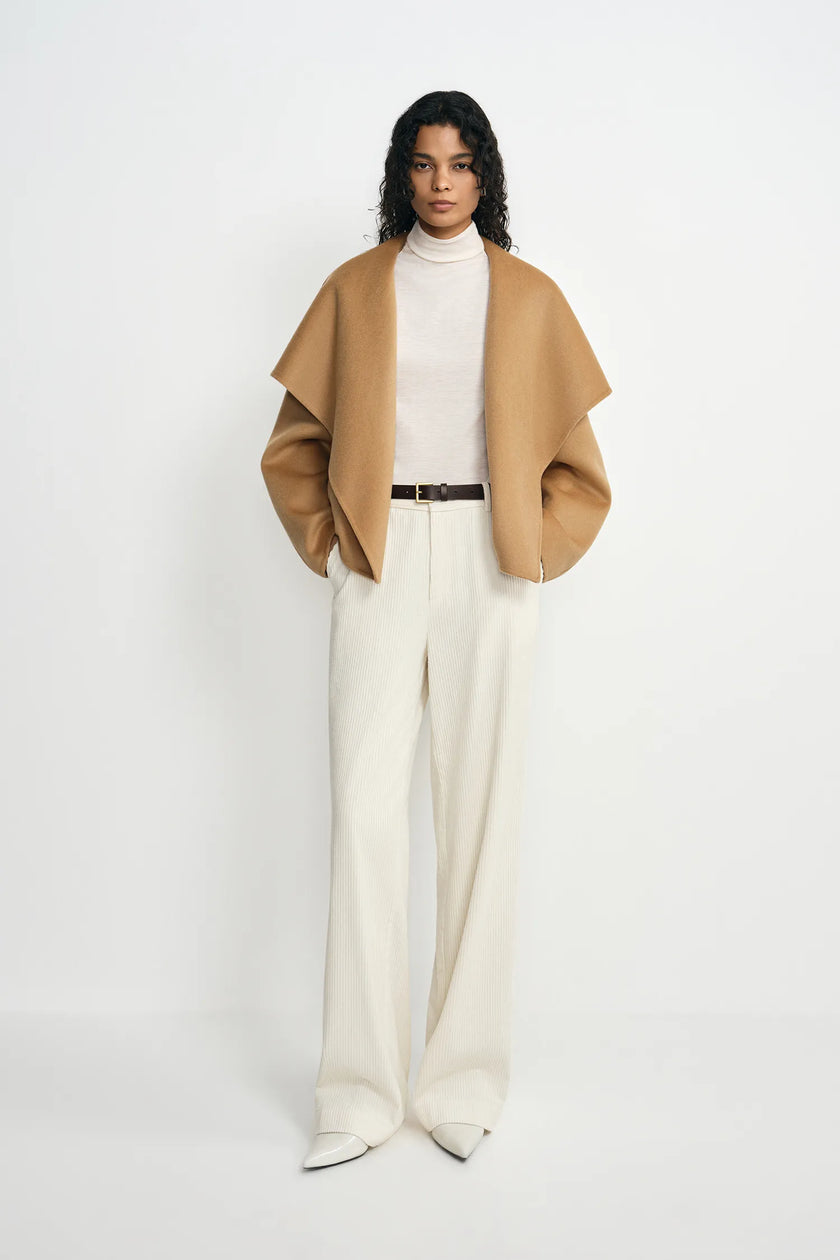 A model stands against a plain white studio background wearing a camel-colored draped cape jacket over a cream turtleneck, paired with cream wide-leg trousers and a thin dark belt.