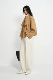 A model stands in a studio wearing a camel wrap-style coat over a white top and off-white wide-leg trousers, holding a black rectangular bag against a plain light background.