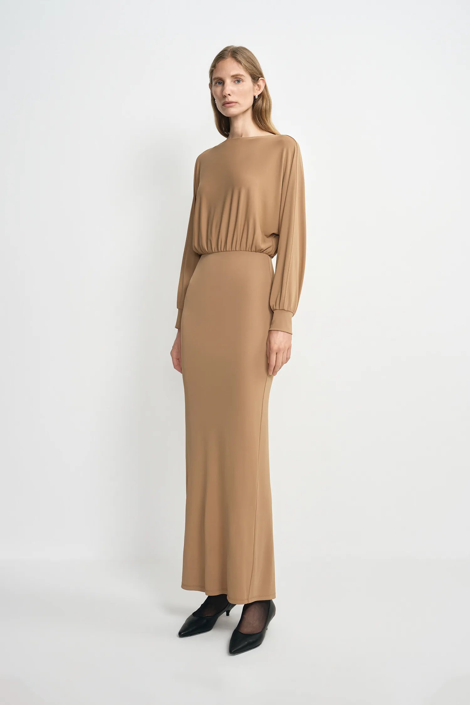 A model wearing a camel soft knit maxi dress with relaxed dolman sleeves and a fluid, ankle-length skirt, pairing minimal black heels.
