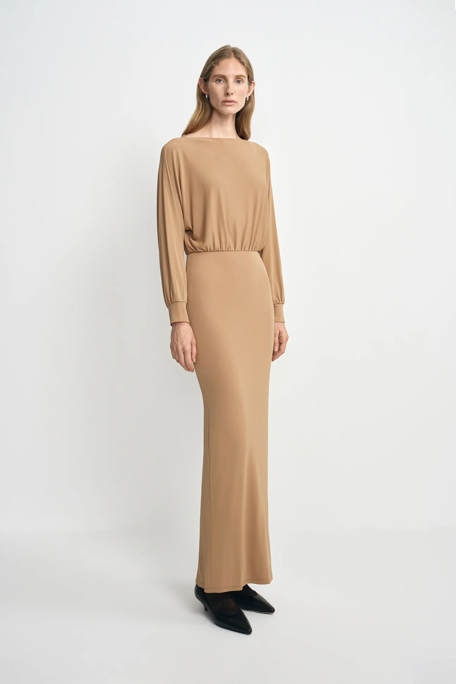 A model wearing a camel soft knit maxi dress with a draped dolman-sleeve top and slim-fitting skirt, pairing classic black pointed-toe heels.